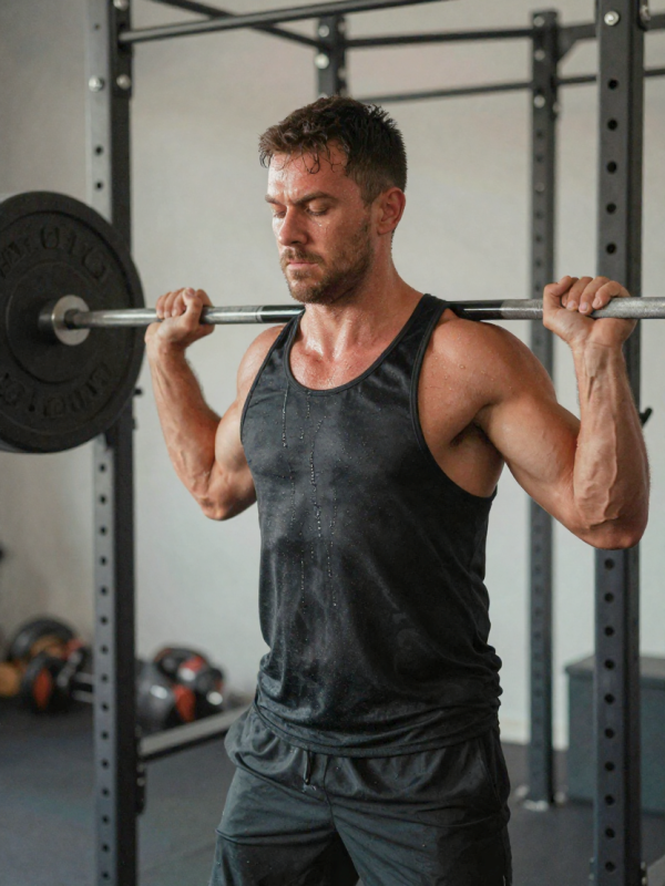 Gespierde man in sportkleding die zijn borstspieren traint met een zware halter, zweet, aderen, fitnessapparatuur op de achtergrond.