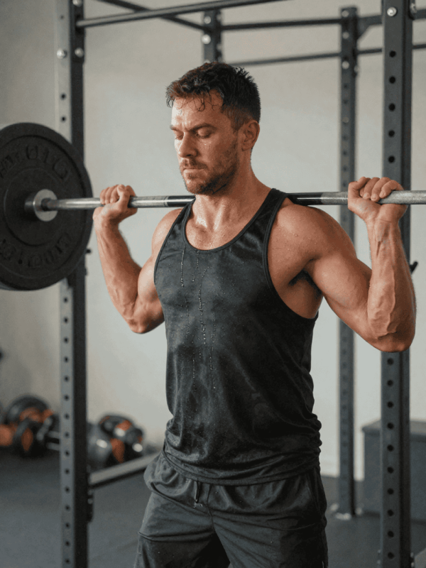 Gespierde man in sportkleding die zijn borstspieren traint met een zware halter, zweet, aderen, fitnessapparatuur op de achtergrond.